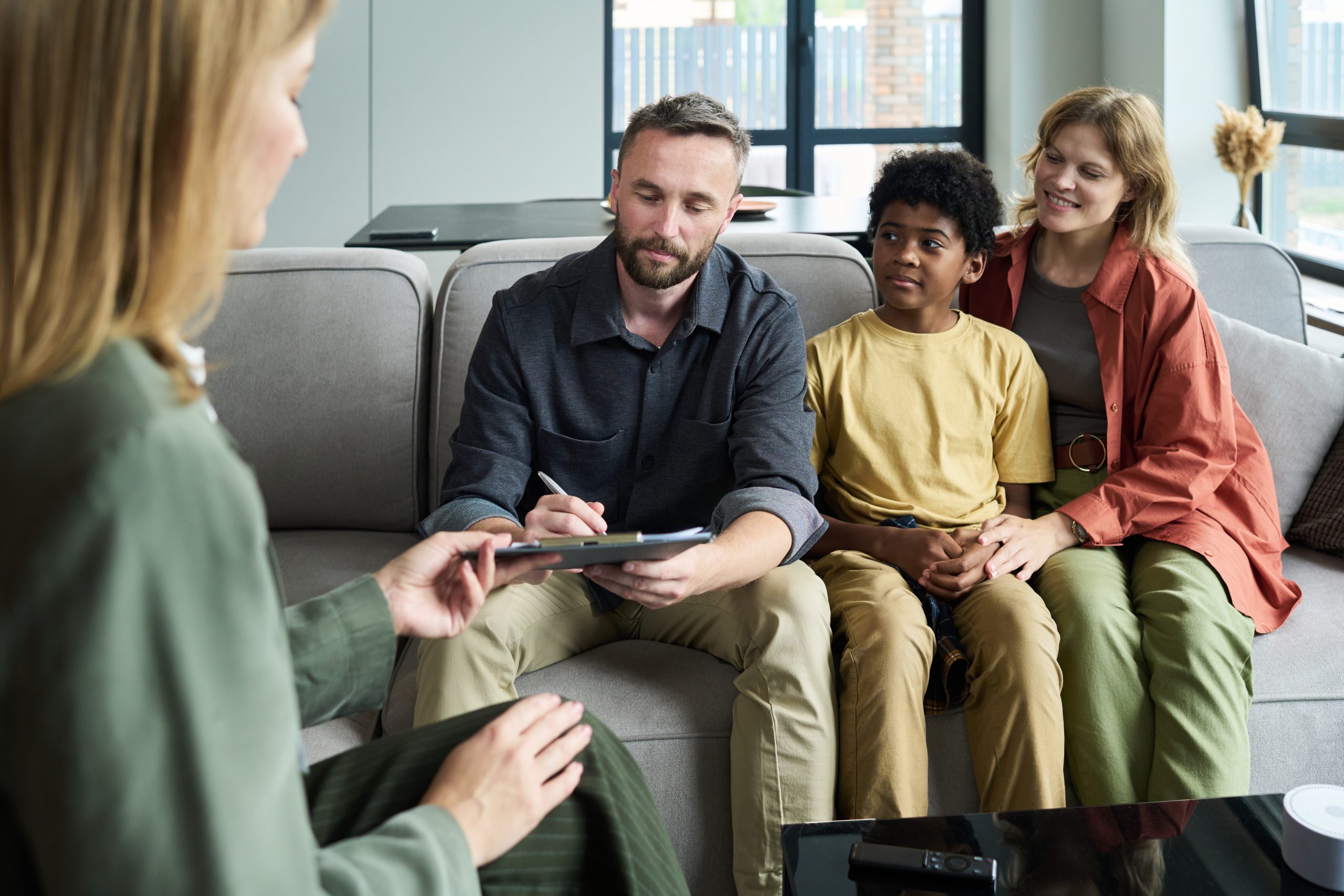 Therapist engaging with family during counseling session at cozy, welcoming office with natural decor. Family includes man, woman, and child sitting on sofa, talking