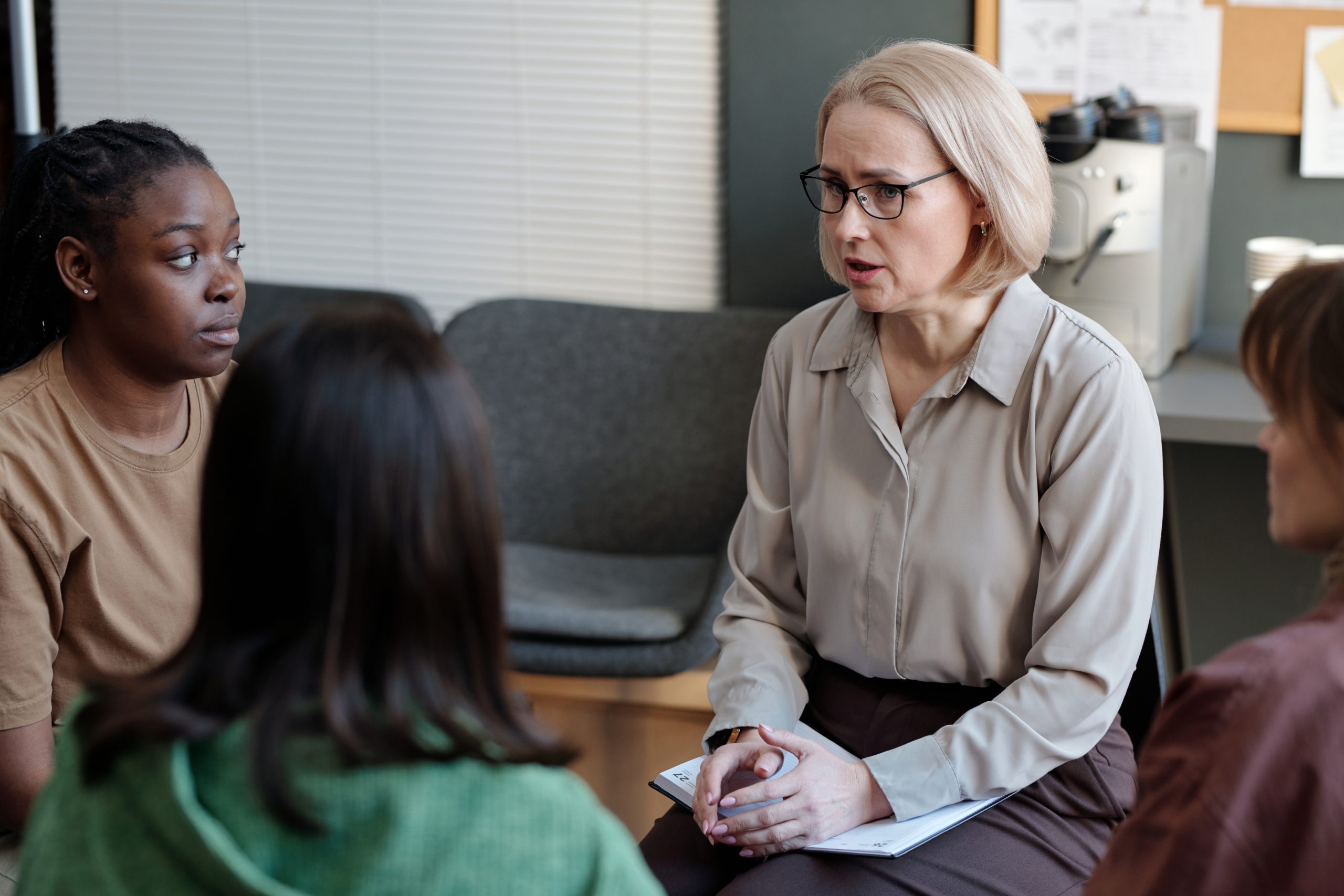 Blond mature female psychologist giving advice to one of two intercultural patients sharing their stories with the rest of session group